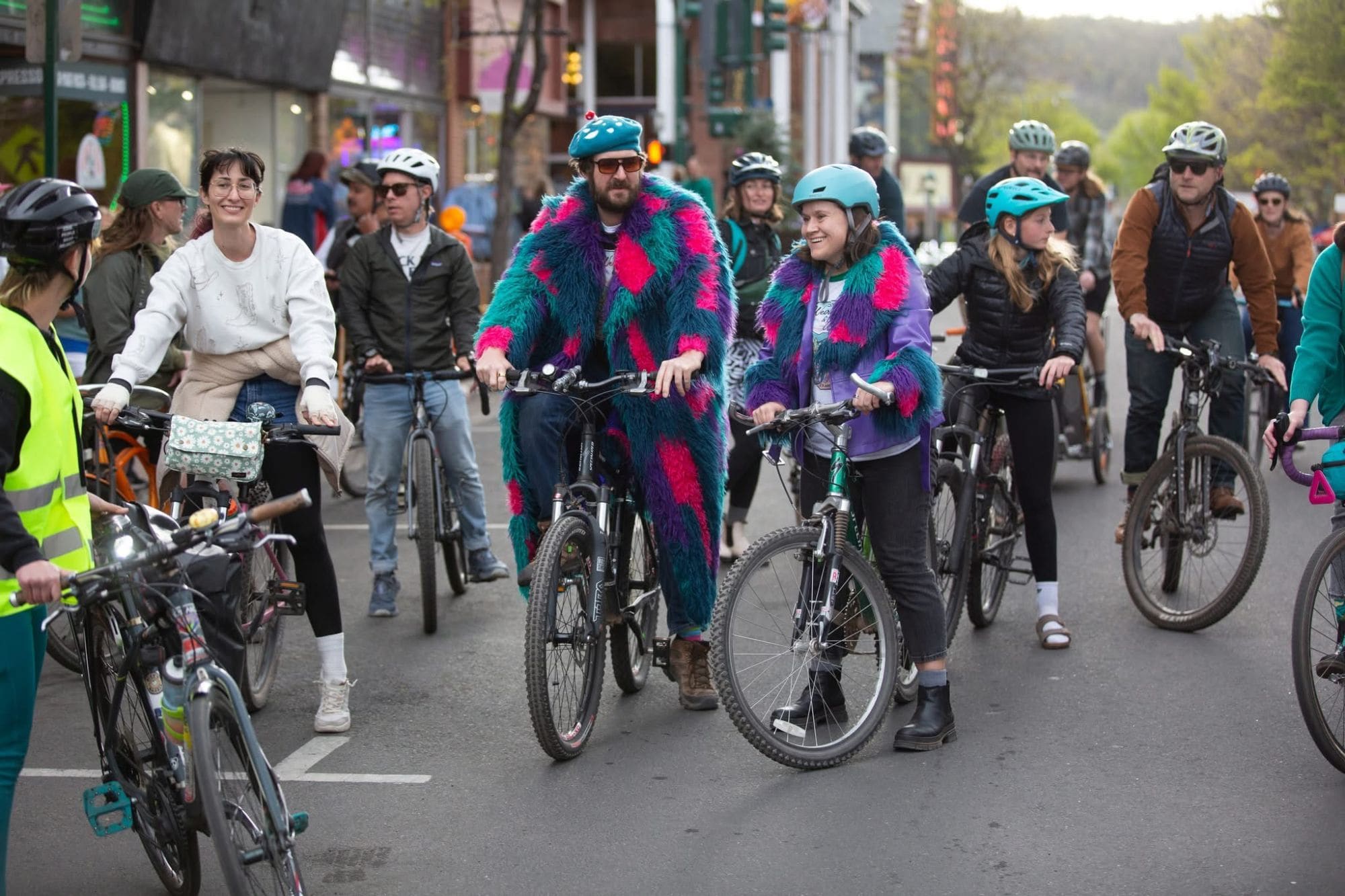 A crowd of riders on bikes filling a car-free downtown Flagstaff street on a First Friday evening, with Nick in the teal, pink and purple shag coat near the front.
