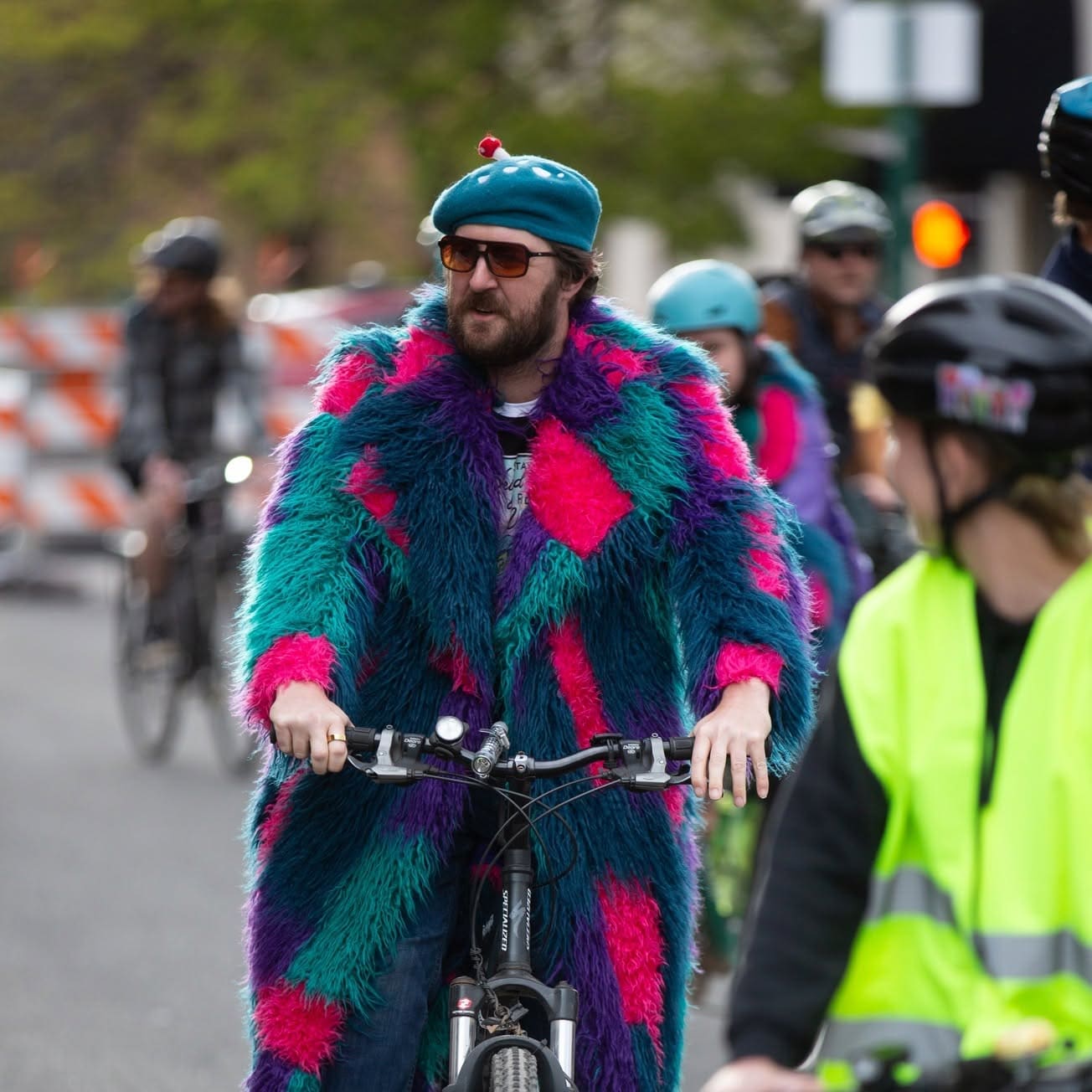 Nick Jones riding a bike in a teal, hot pink and purple shag coat and a teal beret, sunglasses on, in downtown Flagstaff.